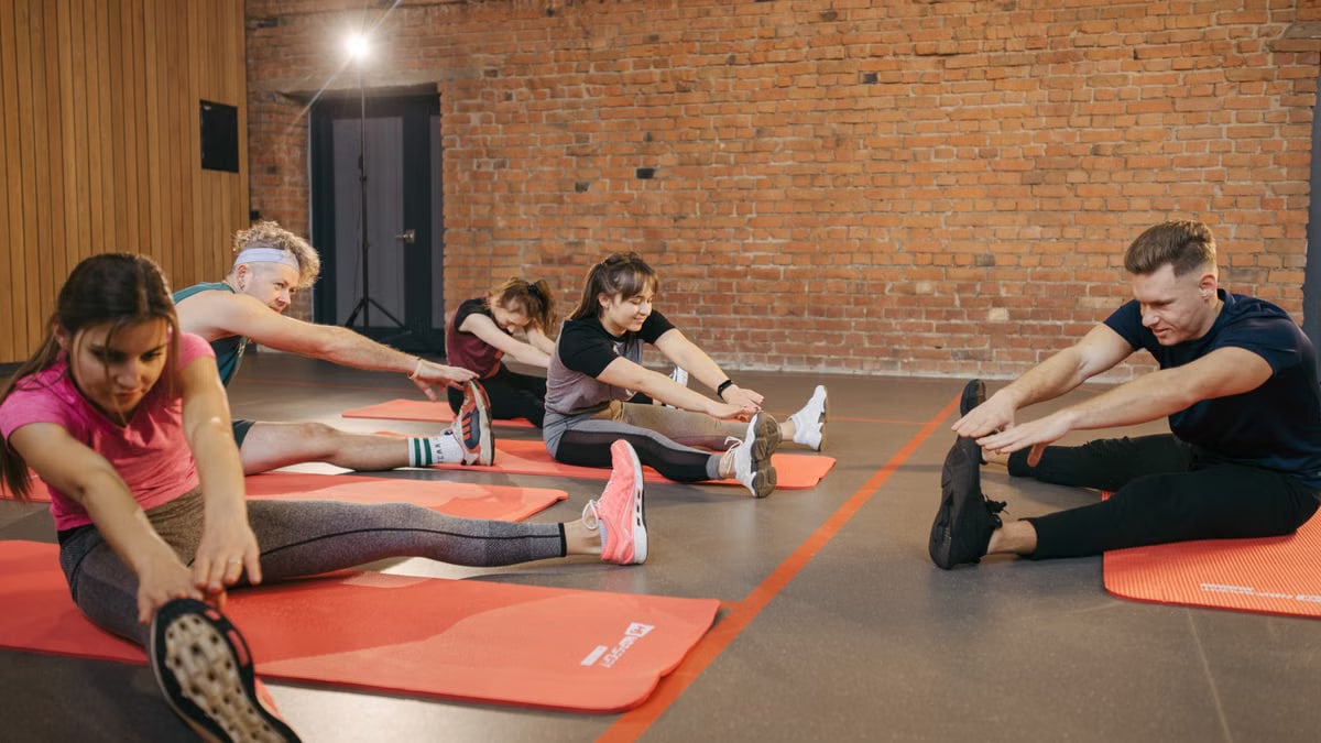 A group of diverse adults stretching on mats in a brick-walled fitness studio.